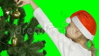 戴着红色圣诞帽的年轻女孩用<strong>金色球</strong>体装饰绿色圣诞树。 准备庆祝一下